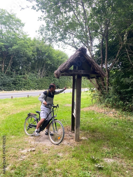 Obraz Middle-aged man on a bicycle stopping in front of an information map at the entrance of a forest, preparing for a nature ride