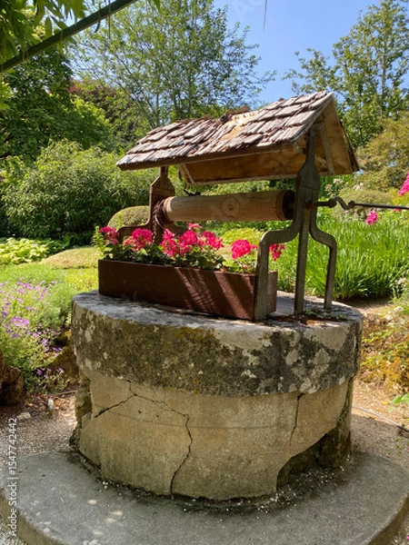 Obraz Old stone well with a small wooden roof and a flower box of red geraniums on the edge, evoking rustic charm and countryside life