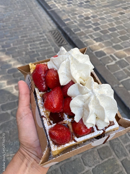Obraz Close-up of a person holding a tray with a Belgian waffle topped with whipped cream, strawberries, and chocolate sauce on a city street.