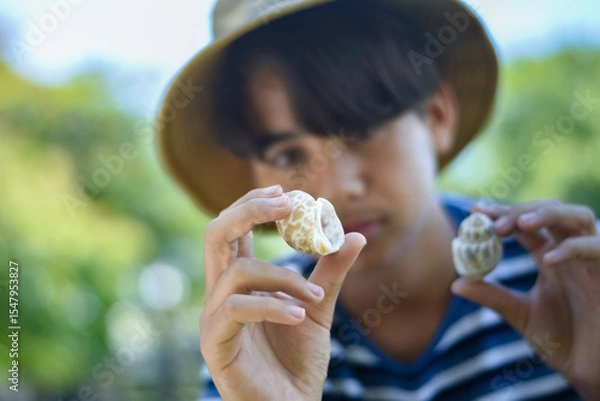 Fototapeta A thoughtful boy closely observes two seashells in his hands. The image reflects curiosity, learning, and a love for nature in an outdoor environment.