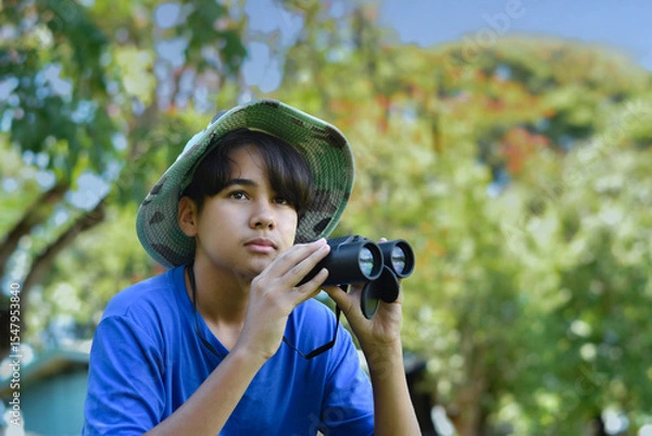Fototapeta A curious boy in a wide-brim hat looks through binoculars during an outdoor exploration. Surrounded by trees, he enjoys a nature adventure on a sunny day.