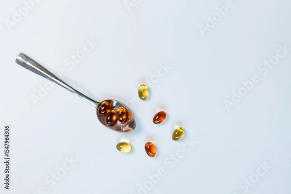 Obraz A close-up, top-view image of golden fish oil capsules on a silver spoon, isolated on a clean white background. This flat lay composition represents the concept of daily nutritional supplements.