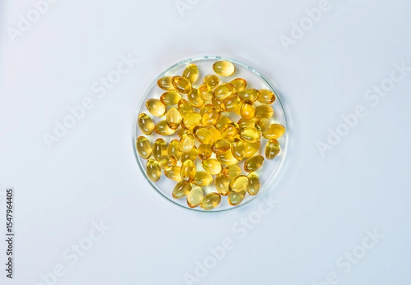 Obraz Top-view of yellow and amber supplement capsules in a glass petri dish, isolated on a white background. This shows scientific research, pharmaceutical development for health products like fish oi.
