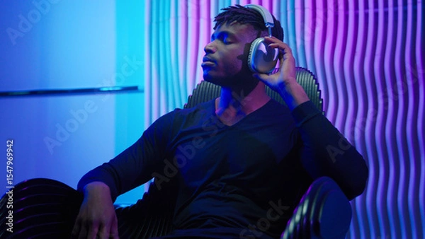 Fototapeta African American man guy male sitting in chair listening music in headphones using wireless earphones listen audio enjoy song sound enjoy modern cyber technology neon futurism room futuristic violet