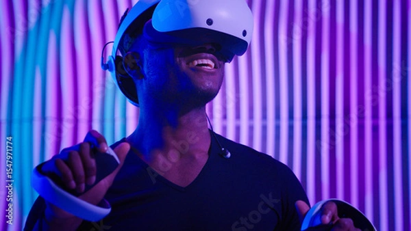 Fototapeta Excited African American man guy male gamer gaming vr play virtual reality glasses helmet internet game modern cyber technology neon futurism indoors futuristic metaverse playing software AR designer
