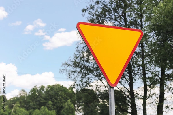 Fototapeta Bright yellow traffic sign. Triangular road warning symbol. Urban infrastructure with trees. Give way triangle sign. Safety signal in countryside. Driving rules and signage visibility reminder.