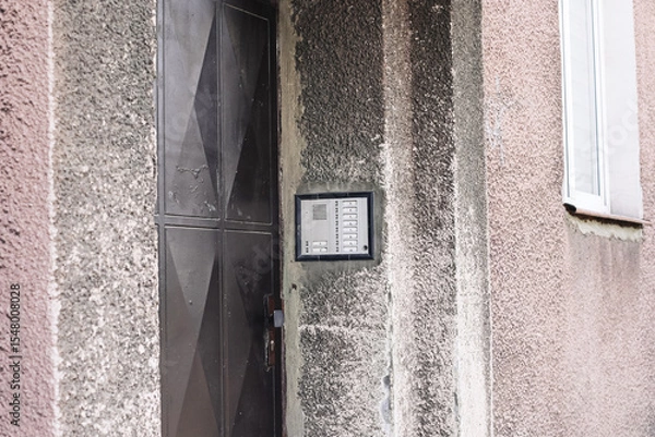 Fototapeta Old apartment intercom system. Entry phone with buttons on weathered wall. Vintage access control panel near metal door. Residential building entrance with textured concrete and worn facade.