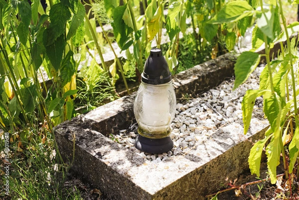 Fototapeta Glass candle on old grave. Cemetery lantern without flame among green plants. Peaceful burial place in summer light. Symbol of memory and silence. Nature surrounding forgotten resting spot.