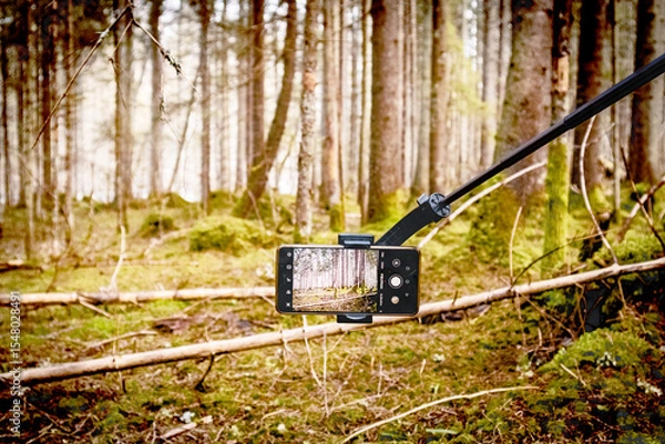 Fototapeta 
A smartphone attached to a selfie stick taking a photo of a forest with trees and mossy ground.