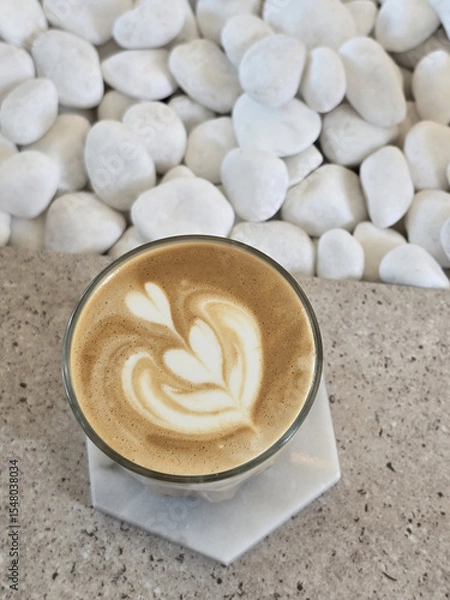 Obraz beautifully crafted latte with intricate foam art sits on marble surface, surrounded by smooth white pebbles, creating serene and inviting atmosphere