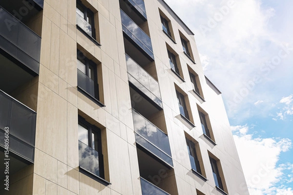 Fototapeta Modern apartment building facade. Residential architecture with balconies and large windows. Urban housing complex under blue sky. Minimalist exterior with beige and black wall materials.