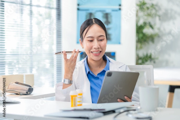 Fototapeta Portrait of an Asian female doctor using a smartphone in a hospital corridor, interacting with a virtual assistant or AI chatbot. She reviews health data via smart mirror technology, embracing future