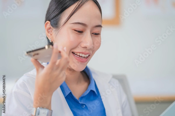 Fototapeta Portrait of an Asian female doctor using a smartphone in a hospital corridor, interacting with a virtual assistant or AI chatbot. She reviews health data via smart mirror technology, embracing future