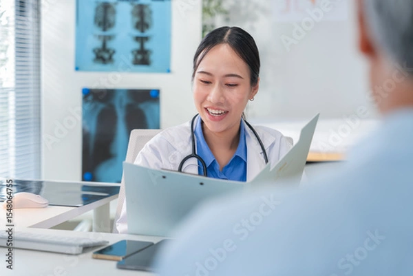 Fototapeta Asian doctor in uniform attentively assists a senior couple with medical paperwork in a hospital area, explaining health records and test results with care and professionalis senior health