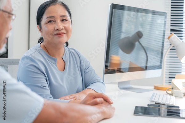 Fototapeta Asian doctor in uniform attentively assists a senior couple with medical paperwork in a hospital area, explaining health records and test results with care and professionalis senior health