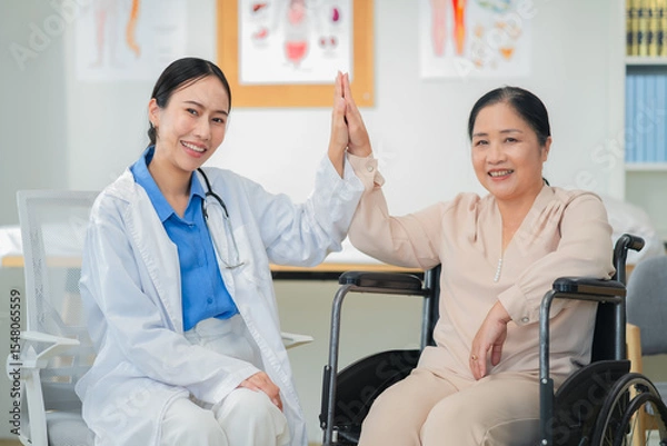 Fototapeta Asian female doctor giving a cheerful high five to an elderly woman patient, both smiling warmly in a medical clinic setting. A joyful moment of healthcare connection, trust, and support between careg