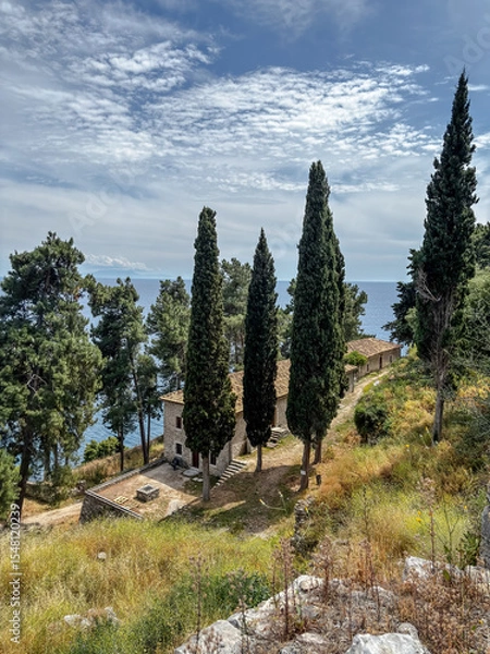 Fototapeta A peaceful stone building surrounded by tall cypress trees on a grassy hillside above the Ionian Sea in Parga, Greece.