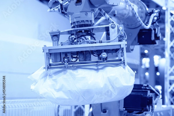 Fototapeta An industrial robotic arm with a pneumatic gripper lifts a heavy white bag in a factory setting. The background shows a manufacturing environment
