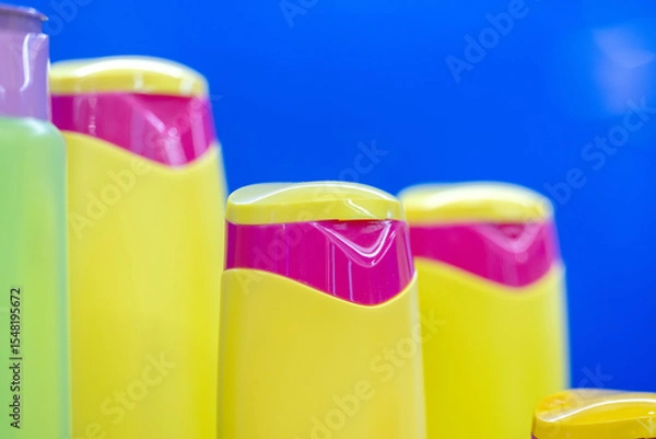 Fototapeta Brightly colored yellow and pink shampoo bottles in a row against a vibrant blue background, showcasing modern packaging design and color contrast, close-up view. Selected focus