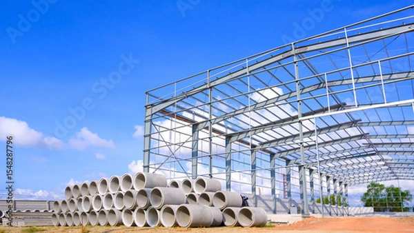 Fototapeta Metal structure of warehouse industrial factory building with concrete drainage pipes stacked on the ground in construction site against blue sky background, perspective side view 
