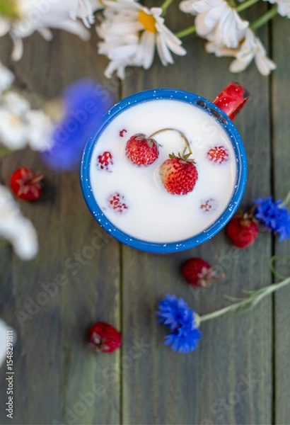 Fototapeta Wild strawberries floating in a blue enamel mug full of milk, placed on a wooden table decorated with daisies and cornflowers, creating a rustic and idyllic breakfast scene