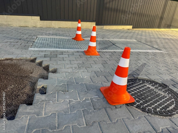 Fototapeta Orange traffic cones mark off a section of uneven pavement near a grate. The bright cones stand out against the gray stones on a sunny afternoon in an urban setting