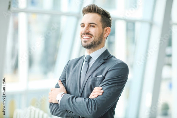 Obraz Confident businessman in suit with folded arms, smiling in a modern office space, exuding professionalism and success. Bright natural light and sleek interior design enhance the ambiance.