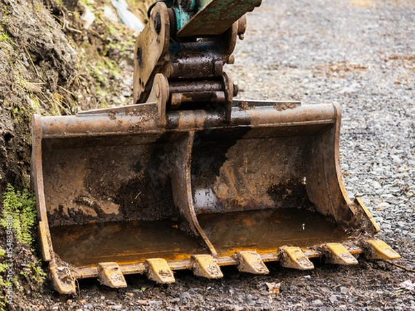Fototapeta Excavator bucket on the ground. Stone pebble on the ground, soil in the background. Park or house construction work concept. Nobody. Heavy machinery tools and equipment.