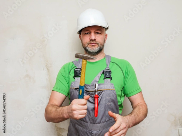 Fototapeta A workman in a green shirt and gray overalls is holding tools Portrait of a male general laborer on a construction site in white safety helmet. Model in his 40s with grey beard.