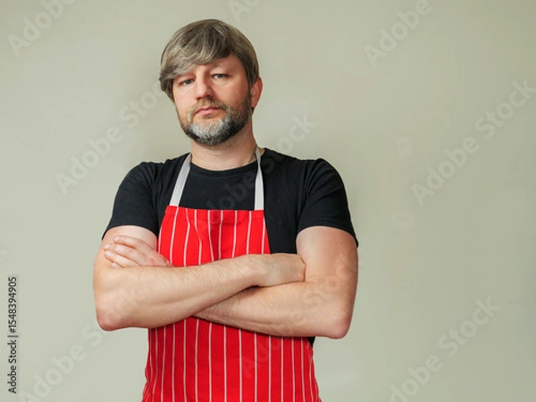 Fototapeta Portrait of professional butcher or fishmonger in black shirt and red and white classic traditional apron, light color background. Male model in his 40s with grey beard and hair. Hands crossed