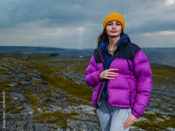 Fototapeta A girl is wearing a purple jacket and a yellow hat stands on a rocky hill overlooking the ocean. The scene is serene and peaceful, with the woman posing for a photo. Burren area, Ireland. Travel 