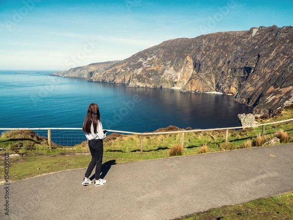 Fototapeta Teenager girl sightseeing stunning Slieve League cliff nature scenery and Atlantic ocean. Blue cloudy sky. County Donegal, Ireland. Unique landmark and tourist point of view and hiking area.