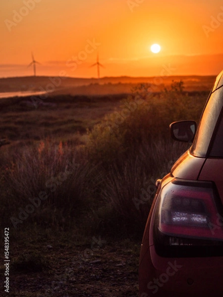 Fototapeta A red car is parked in a field with wind turbines and a warm sunrise in the background. The car is facing the sun, and the sky is orange and pink. The scene is peaceful and serene. Early bird concept