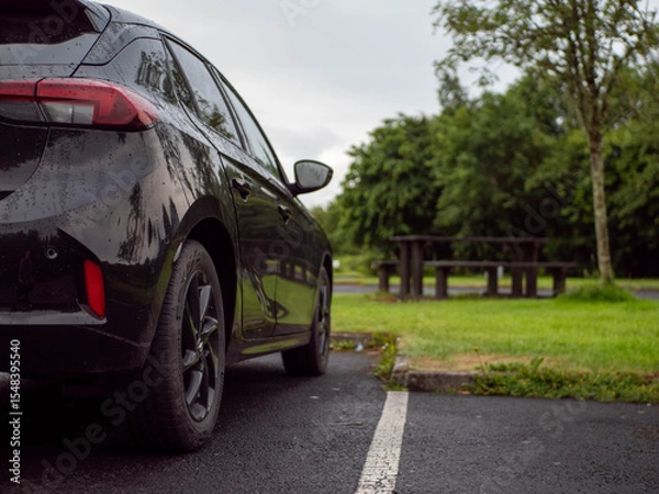 Fototapeta Black family car parked in a parking lot of a park with barbecue or dining area, selective focus. Nobody. Car surface is with rain drops. Outdoor activity. Travel and tourism.