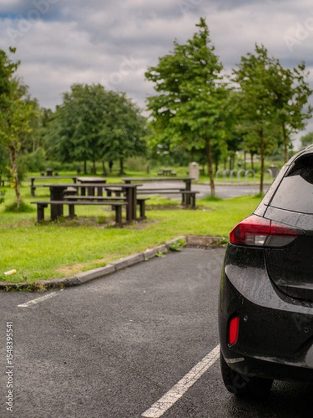Fototapeta Black family car parked in a parking lot of a park with barbecue or dining area, selective focus. Nobody. Car surface is with rain drops. Outdoor activity. Travel and tourism.