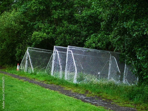 Fototapeta Football or soccer goal posts stacked off training pitch by dark trees. Professional club training field and equipment. World popular sport.