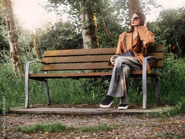 Fototapeta Young and stylish teenager girl sitting on a bench in a park alone. Waiting for date concept. The model wears trendy brown jacket and dark glasses and looks calm and elegant. Selective focus.