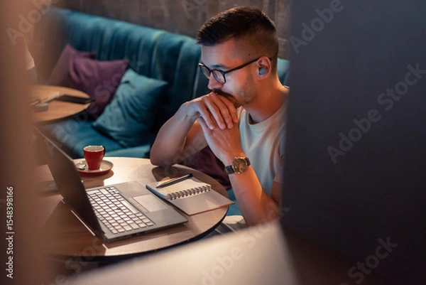 Fototapeta Focused young man wearing glasses and wireless earbuds, watching a webinar, having an online course on a laptop in a modern interior. 