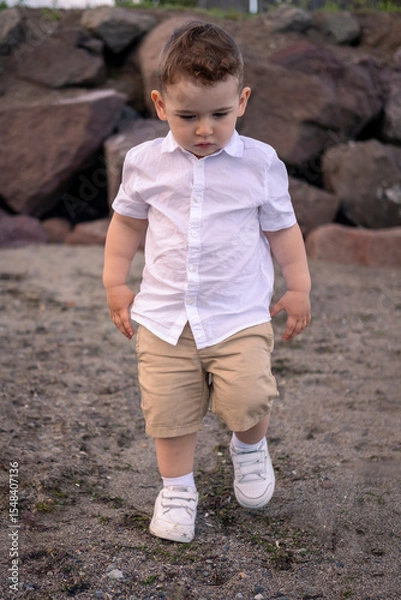 Fototapeta Child enjoying a quiet beach afternoon. little boy walking on the beach. Curious boy in white shirt walks along the shore.