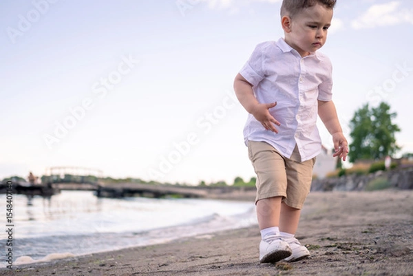 Fototapeta young boy running on the beach. Child enjoying a quiet beach afternoon. little boy walking on the beach. Curious boy in white shirt walks along the shore.