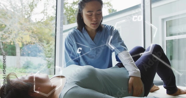 Obraz Physiotherapist in scrubs raising patient's bent leg on table in clinic by windows with AR overlays