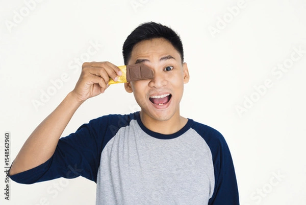 Fototapeta Young man holding golden-wrapped chocolate bar over his eye, expressing playful enthusiasm. Ideal image for themes of happiness, fun, sweets, and youthful enjoyment.