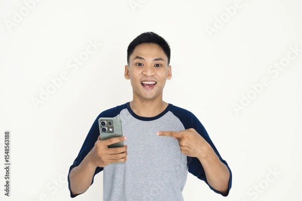 Fototapeta A cheerful young man wearing a casual shirt, joyfully holding a smartphone and pointing to it, expressing amazement.