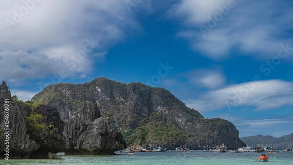 Fototapeta Many Filipino bangka boats are anchored in the turquoise lagoon. A tourist canoe is sailing. Picturesque cliffs with steep slopes and green vegetation against a blue sky and clouds. Philippines.