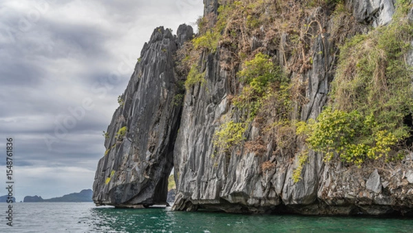 Fototapeta A picturesque cliff in the ocean. There is green tropical vegetation on the steep, furrowed slopes. The gap between the rocks is visible. Clouds in the sky. Philippines. Palawan. El Nido. 