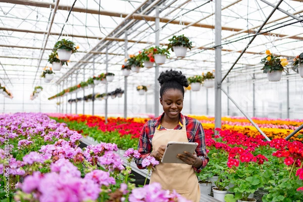 Obraz An African American woman smiles while using a tablet in a colorful flower greenhouse, surrounded by blooming plants.