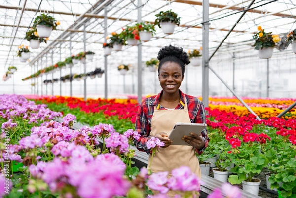 Obraz A smiling African woman in a greenhouse with a tablet, surrounded by vibrant flowers and plants, and other potted plants.