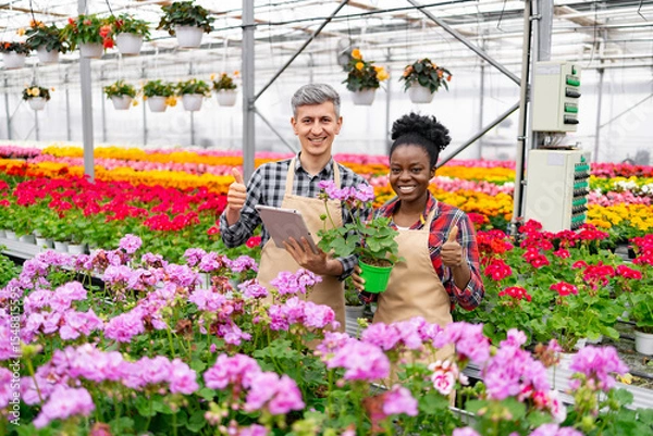 Obraz Two smiling people in a greenhouse with flowers, giving thumbs up. One holds a tablet, the other a plant.