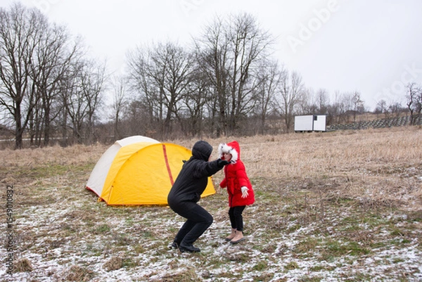 Fototapeta Joyful moment in winter wilderness as child in red coat plays with adult near cozy yellow tent under cloudy skies