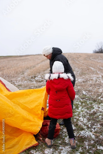 Fototapeta Preparing for a winter adventure as a child and adult set up a bright yellow tent in a snowy field under overcast skies